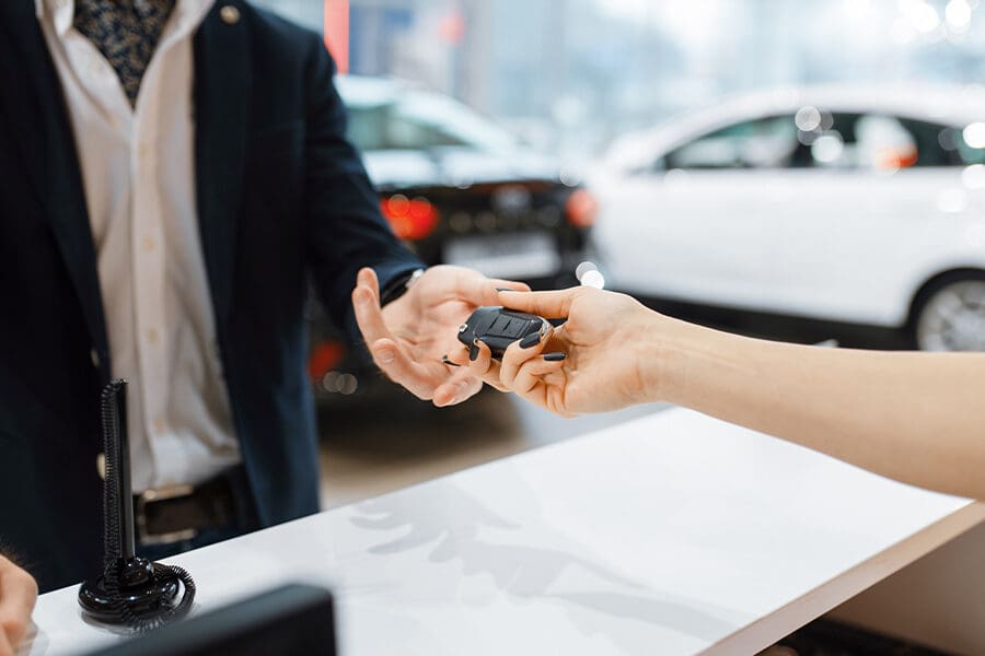 Should I Purchase Rental Car Insurance? - Man Takes the Key From New Auto in Car Dealership as the Appers Are Being Signed a the Sun is Shining on the New Cars