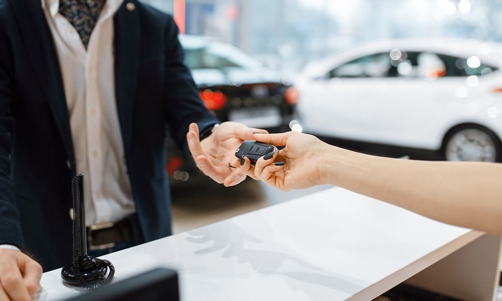 Should I Purchase Rental Car Insurance? - Man Takes the Key From New Auto in Car Dealership as the Appers Are Being Signed a the Sun is Shining on the New Cars